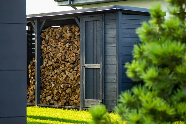 Modern siyah ahşap bir barınağın altına düzgünce istiflenmiş yakacak odun, yeşil çimenler ve ön planda bulanık bir çam ağacı. Kırsal sakinlik, modern bir dokunuş..