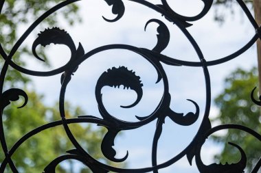 Çiçek desenli süslü demir bir çitin yakın plan yeşil ağaçların ve mavi gökyüzünün bulanık zeminine karşı.