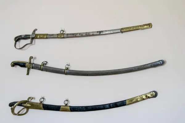 Süslü kabzalı ve kılıç kınalı üç antika kılıç beyaz arka planda yatay olarak sergileniyor. Pirinç ve metal işlemeli tarihi askeri kılıçlar..