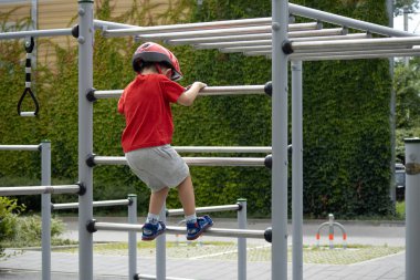 Çocuk dışarıda oyun bahçesinde maymuna tırmanırken kırmızı tişört ve kask giyiyor. Aktif çocukluk ve fiziksel gelişim kavramı.