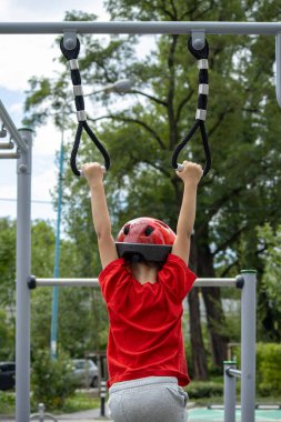Kırmızı tişörtlü ve kasklı bir çocuk açık hava çocuk parkında jimnastik yüzüklerine asılmış. Şehir parkındaki çocuklar için aktif oyun ve spor.