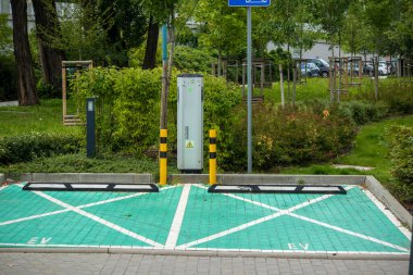 Boş elektrikli araç, modern bir şehir parkı bölgesinde yeşil kaldırım ve elektrik santraliyle dolu park yerlerini dolduruyor..