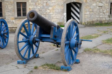 Mavi ahşap tekerleklerdeki eski siyah top taş kale duvarının yanında sergileniyordu. Geçmiş yüzyıllardan kalma tarihi askeri topçu parçası..