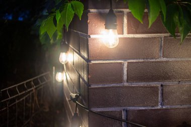 İp lambaları geceleri tuğla bir duvar boyunca asılı durur, sıcak bir parıltı yayar. Görüntünün üstünü çerçeveler. Huzurlu, rahat bir akşam manzarası..