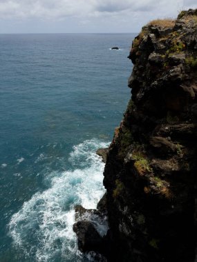 Dikey çekim. Okyanus dalgaları Portekiz 'in Madeira sahilindeki kayalık kıyı şeridine çarpıyor. Beyaz sörf spreyli deniz pelerini ve engebeli taşlar.