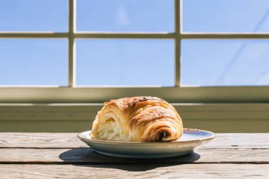 Lezzetli bir altın kruvasan, parlak mavi gökyüzü ve beyaz camları olan bir pencerenin önündeki dekoratif bir tabakta oturur..