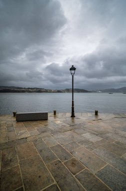 Huzurlu körfez manzarası, içinde lamba direği olan taştan set manzarası ve dramatik bulutlu gökyüzünün altında bank.