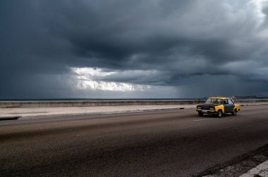 Havana, Küba, 19 Ağustos 2023: Havana, Küba 'da, dramatik, karanlık fırtına bulutları altında Malecon üzerinde sürüş sarı taksi