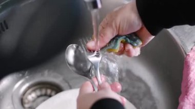 Çatal bıçakları dikkatlice yıkayan kadın musluğun altında sarı temizlik süngeri ile ev lavabosunda detaylı mutfak temizliği yapıyor.