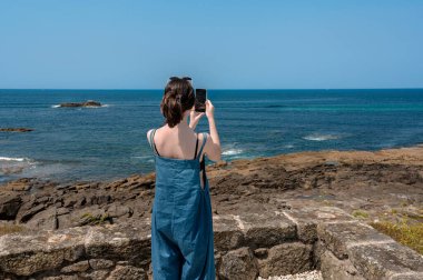 Genç bir kadın turist, parlak ve güneşli bir yaz gününde akıllı telefonuyla büyüleyici deniz manzaralarını yakalıyor. Okyanusun güzelliğinin ve sahil şeridinin huzurunun tadını çıkarıyor.