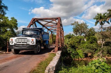 Eski ulaşım kamyonu tropikal bir kırsal bölgede nehrin üzerinden paslı metalik bir köprüyü geçiyor.