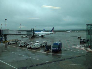 Bulutlu bir günde havaalanı pisti manzarası, park halindeki uçaklar ve yer servis araçları. Sahne ıslak, yakın zamanda yağmur yağdığını gösteriyor, ön planda modern bir terminal var..