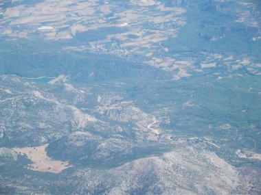 Yeşil vadileri ve tarımsal alanları olan dağlık bir arazinin havadan görünüşü. Arazide kayalık tepeler ve ekili arazi parçaları yer alıyor ve doğal ve insan tarafından değiştirilmiş ortamların bir karışımını sergiliyor..