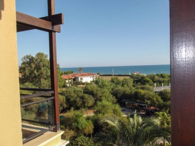 Yemyeşil bir manzaraya ve okyanusa bakan bir balkondan manzara. Gökyüzü birkaç bulutla berrak ve su canlı bir mavidir. Yakın yapılar görülebilir, ağaçlarla çevrili..