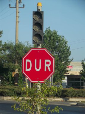 Kırmızı dur işareti, üzerinde beyaz harflerle 'DUR' yazıyor, trafik ışıklarının yanındaki direğe monte edilmiş. Arka planda ağaçlar ve açık mavi gökyüzü olan bir bina yer alıyor..