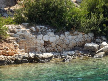 Berrak turkuaz su ve yemyeşil verimli bir kıyı şeridi. Taş duvar yıpranmış ve kısmen batırılmış görünüyor. Bu da kıyıya yakın antik bir yapıyı işaret ediyor..