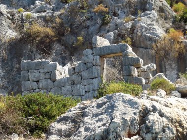 Eski taş kalıntıları kısmen bitki örtüsüyle kaplı, kayalık bir uçuruma karşı kurulmuş. Yapıda büyük, düzensiz taşlar ve kuru çalılar ve yeşilliklerle çevrili açık bir kapı var..