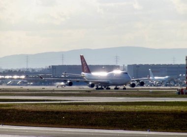 Büyük bir uçak, muhtemelen bir Boeing 747, diğer uçak ve binaların bulanık arka planına sahip bir havaalanı pistinde taksicilik yapıyor..