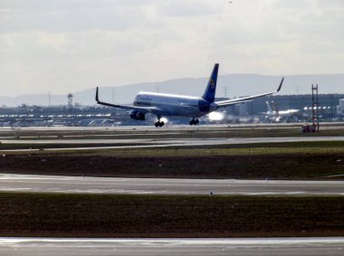 Arka planda bulutlu bir gökyüzü olan ticari bir uçak podyuma iniyor. Uçak, kuyruğunu ve kanatlarını gösteren bir açıyla konumlandırıldı..