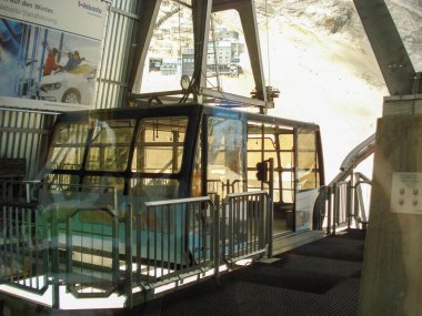 Dağlık bir bölgede cam bir kulübesi olan modern bir teleferik istasyonu. İstasyonda metal yapılar ve çevreleyen manzaralar yer alıyor..