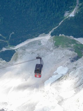 Dağ zirvesinden inen, kayalık arazi ve yeşilliklerle çevrili bir teleferiğin hava görüntüsü. Teleferik kırmızı ve siyah, kablolar mesafeye kadar uzanıyor..