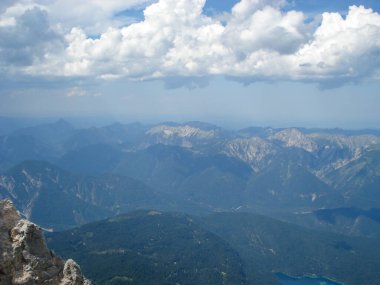 Parçalı bulutlu bir gökyüzünün altındaki engebeli dağların panoramik görüntüsü. Arazide yeşil ormanlar ve kayalık tepelerin karışımı yer alıyor ve uzak mesafeler ufka doğru kayboluyor..