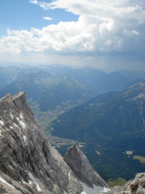 Dağların zirvesinden nefes kesici bir manzara, engebeli kayalık oluşumlar ve aşağıdaki yemyeşil vadiler. Gökyüzü dramatik bulut oluşumlarıyla parçalı bulutlu, manzaranın güzelliğini arttırıyor..