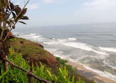 Kıyıda dalgaların dalgalandığı bir kıyı şeridi manzarası. Bereketli yeşillik ve kayalık kayalıklar açık bir gökyüzünün altında sahili çevreledi..