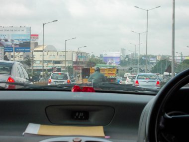 Trafiğin yoğun olduğu bir şehir yolunu gösteren bir arabanın içinden görüntü. Arka planda arabalar, kamyonlar ve reklam panoları gibi çeşitli araçlar var. Gökyüzü bulutlu, bulutlu bir günü işaret ediyor..
