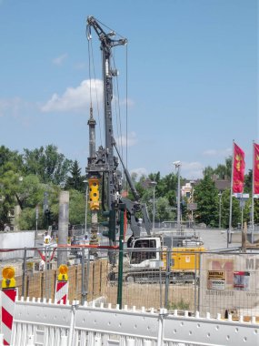 Güvenlik bariyerleri ve inşaat ekipmanlarıyla çevrili büyük bir sondaj makinesinin olduğu bir inşaat alanı. Arkaplanda açık mavi gökyüzü ile ağaçlar ve bayraklar yer alıyor.