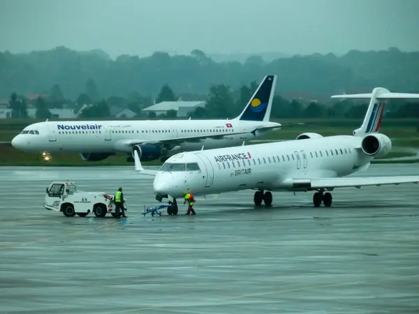 Yağmurlu bir günde havaalanı pistinde iki uçak. Biri Airbus A320, Nouvelair 'de, diğeri de Air France renklerinde bir bombardıman uçağı CRJ-700, yer ekibi kalkışa hazırlanıyor..
