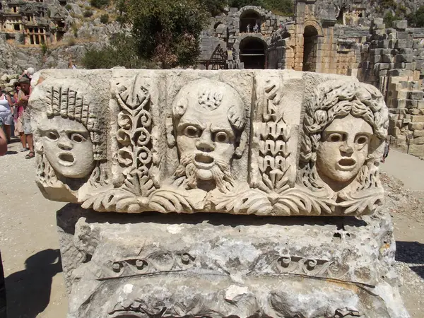 Karmaşık detaylar ve ifadeler içeren, üç oyulmuş yüzün yer aldığı antik bir taş rahatlama. Arkaplanda harabeler ve kayalık bir manzara var. Bu da tarihsel önemi gösteriyor..