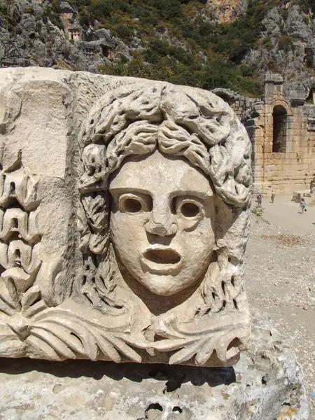 Yapraklarla çevrili karmaşık detaylarla dolu bir yüzü tasvir eden antik bir taşın yakın çekimi. Heykel, kayalık bir zemin ve antik harabelerin üzerine kurulmuş, şaşırmış bir ifadeye sahip bir figürü gösteriyor..