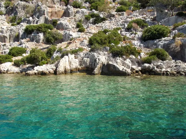 Berrak turkuaz suların kayalık kıyılara çarptığı sakin bir kıyı manzarası. Kayalık arazi yemyeşil çalılarla süslenerek suyla güzel bir kontrast oluşturuyor..