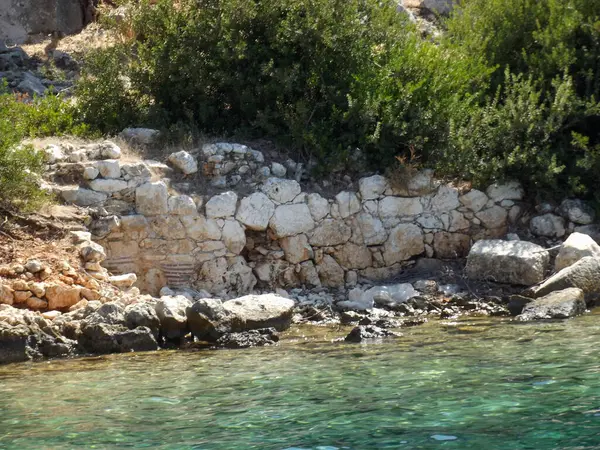 Berrak turkuaz su ve yemyeşil verimli bir kıyı şeridi. Taş duvar yıpranmış ve kısmen batırılmış görünüyor. Bu da kıyıya yakın antik bir yapıyı işaret ediyor..