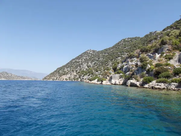 Berrak mavi suların ve yeşilliklerle kaplı kayalık tepelerin olduğu sakin bir kıyı manzarası. Manzara, parlak mavi gökyüzü ile sakin bir atmosfer sergiliyor..