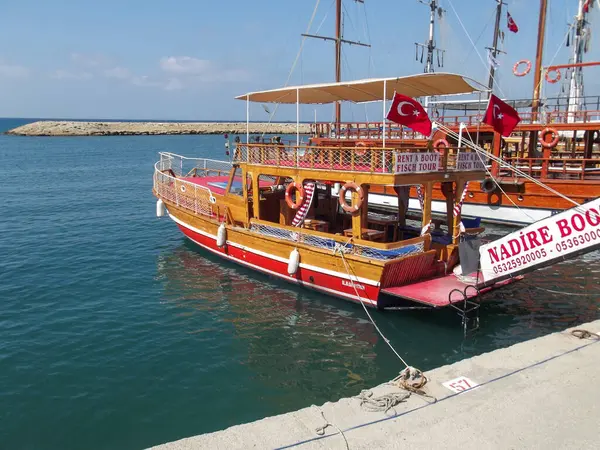 İçinde Türk bayrakları ve tekne kiralama tabelasının bulunduğu bir marinaya demirli ahşap bir tekne. Su sakin ve mavidir, arka planda berrak bir gökyüzü vardır..