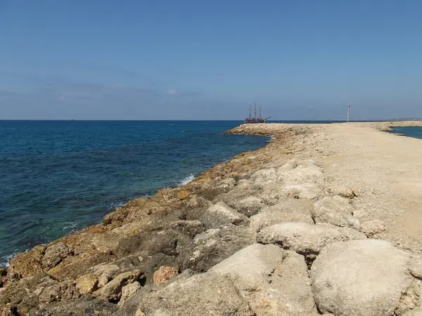 Parlak bir gökyüzünün altında kayalık bir kıyı şeridi açık mavi bir denize uzanıyor. Uzakta, yelkenli bir gemi, sakin manzaraya bir deniz elementi ekleyerek demirler..