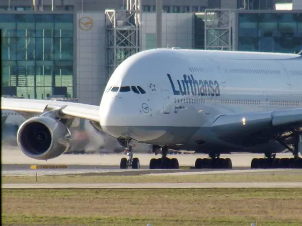 Pistteki bir Lufthansa Airbus A380 uçağı, büyük boyutunu ve kendine özgü giysisini sergiliyor. Arka planda havaalanı binaları ve altyapı yer alıyor.