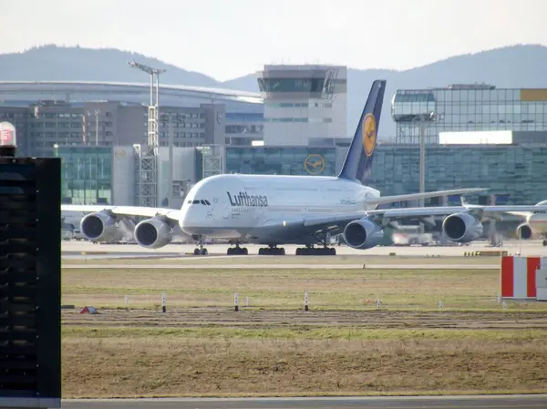 Bir Lufthansa Airbus A380 havaalanında pistte, arka planda modern terminal binaları ve uzak mesafedeki dağlarla taksicilik yapıyor..
