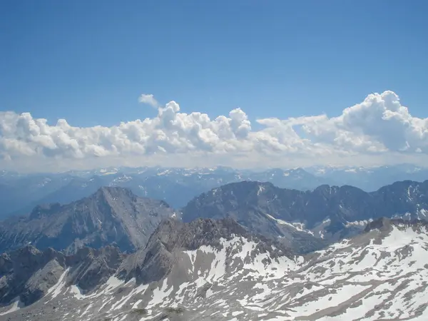 Engebeli tepeler, karla kaplı zirveler ve tüylü beyaz bulutları olan açık mavi bir gökyüzü içeren nefes kesici bir dağ manzarası. Sahne, huzurlu bir ortamda doğanın güzelliğini yakalar..