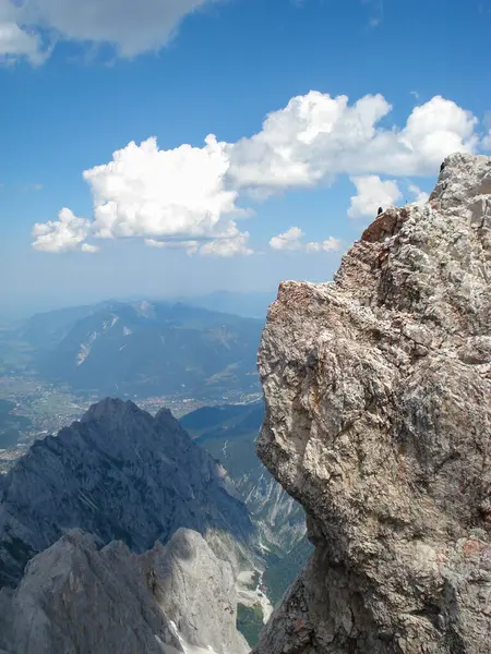 Uçsuz bucaksız bir vadi ve engebeli kaya oluşumlarıyla nefes kesici bir dağ manzarası. Gökyüzü pofuduk beyaz bulutlarla kaplı, dingin bir atmosfer yaratıyor..