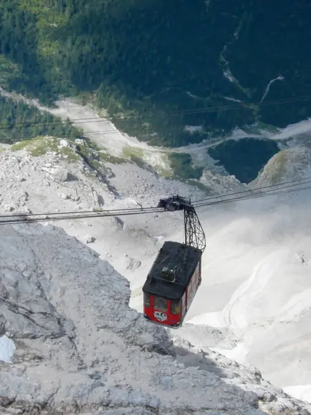Yeşil ve dik uçurumlarla çevrili kayalık bir dağ manzarası üzerinde asılı kırmızı bir teleferik. Teleferik bir tel boyunca seyahat ediyor, aşağıdaki vadiyi nefes kesici bir şekilde gösteriyor..