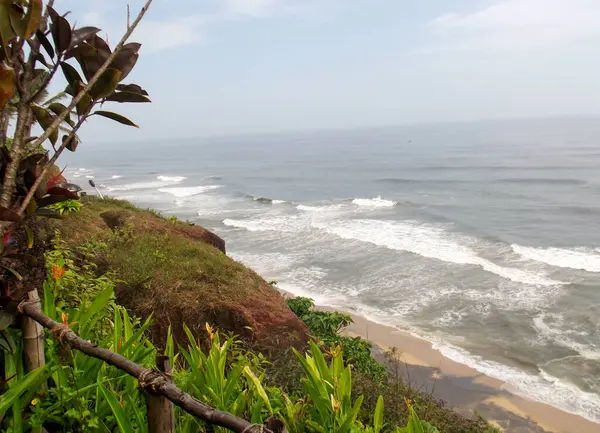 Kıyıda dalgaların dalgalandığı bir kıyı şeridi manzarası. Bereketli yeşillik ve kayalık kayalıklar açık bir gökyüzünün altında sahili çevreledi..