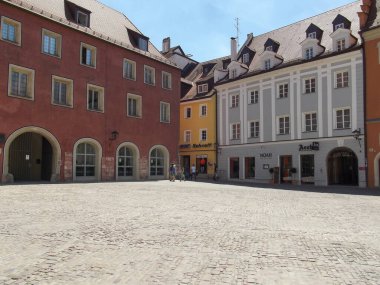 Renkli cepheleri, kaldırımları ve açık mavi gökyüzü olan tarihi binaların yer aldığı pitoresk bir meydan. İnsanlar bölgede rahatça yürüyor..