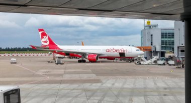 Havaalanı terminaline park etmiş bir uçak. Uçak gövdesinde Airberlin logosu var. Sahnede zemin servis araçları ve arka planda modern bir terminal binası yer alıyor..