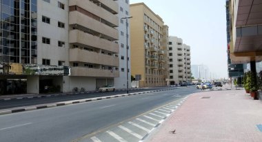 Her iki tarafta da açık mavi gökyüzü olan modern binaların yer aldığı sakin bir şehir manzarası. Yol çoğunlukla boş, birkaç park etmiş araba ve bisiklet sergileniyor. Kaldırımlar yaya ve araç yolları karışımıyla iyi korunmuş..