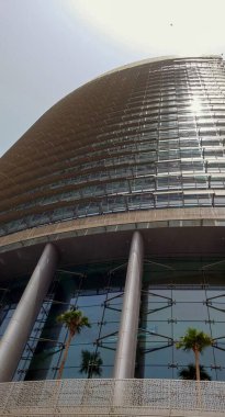 Kıvrımlı cam cepheli modern bir gökdelen, eşsiz bir mimari tasarım sergiliyor. Binada, gökyüzüne karşı dizilmiş büyük sütunlar ve palmiye ağaçları yer alıyor..