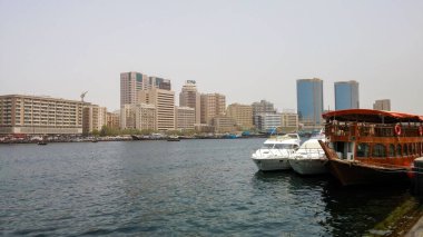 Arka planda modern gökdelenler, kıyıya yanaşmış tekneler ve açık bir gökyüzü olan bir rıhtım manzarası..