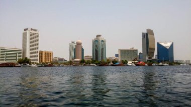 Rıhtım boyunca yüksek binaları olan modern bir şehrin ufuk çizgisi manzarası. Sahne, suya yansıyan cam ve beton yapılar da dahil olmak üzere mimari tarzların bir karışımını yakalıyor..